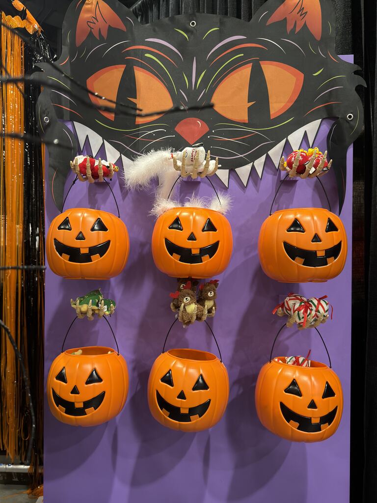 A purple wall display featuring a large cat cutout above six plastic jack-o’-lantern trick-or-treat buckets, each adorned with small plush toys hanging from their handles.