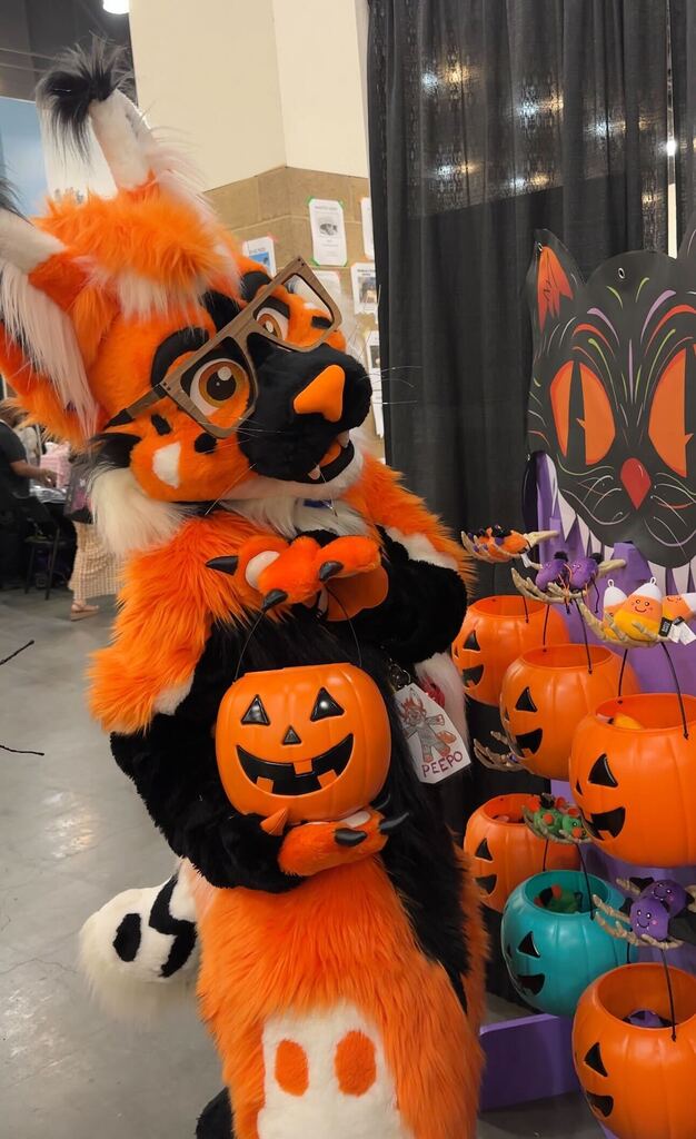 A person in an orange and black fox fursuit with glasses holds a jack-o’-lantern candy bucket next to a display of Halloween-themed treat pails.