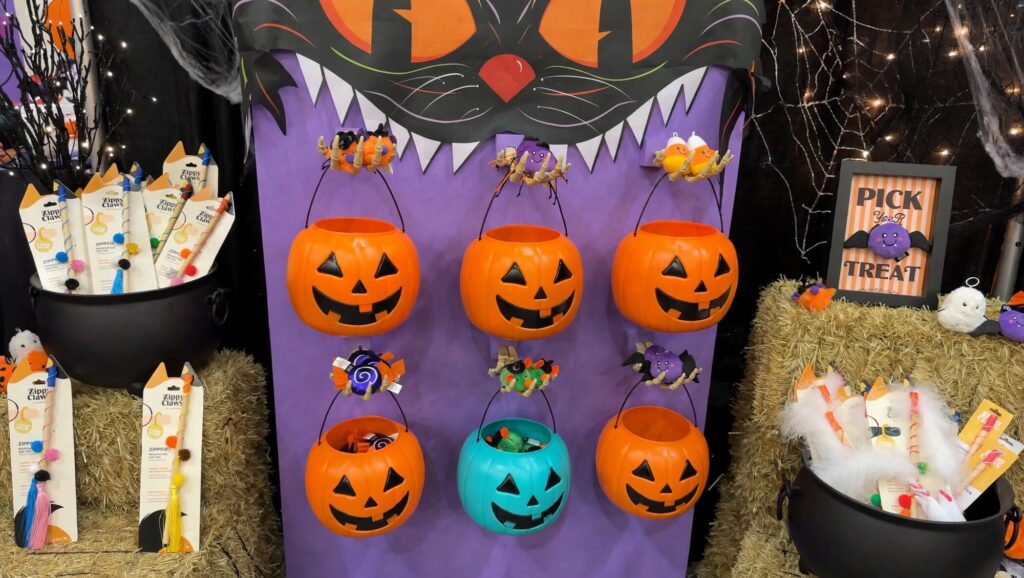 A Halloween display with six jack-o’-lantern buckets hanging on a purple board, flanked by black cauldrons filled with ZippyClaws pet toys and a “Pick Your Treat” sign.